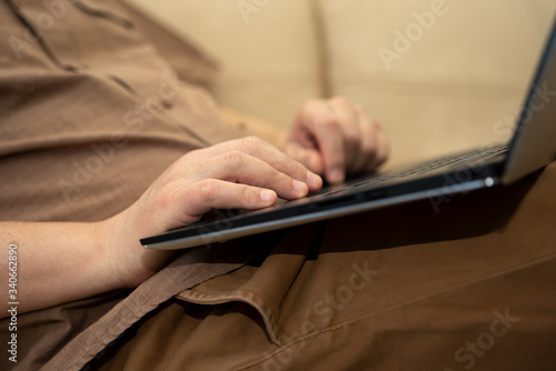 A man reclining on the sofa working on a laptop at home. Close-up side shows: a fragment of the body, hands and laptop. Selective focus.