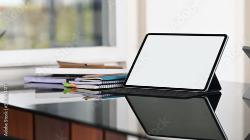 Computer tablet with keyboard case putting together on glass working desk with stack of books and notebook over comfortable workplace as background.