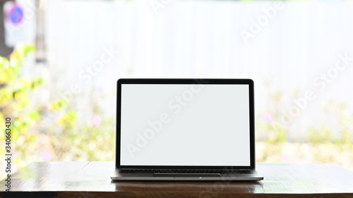 Computer laptop with white blank screen putting on wooden working desk over flowers with sunny outdoors as background.