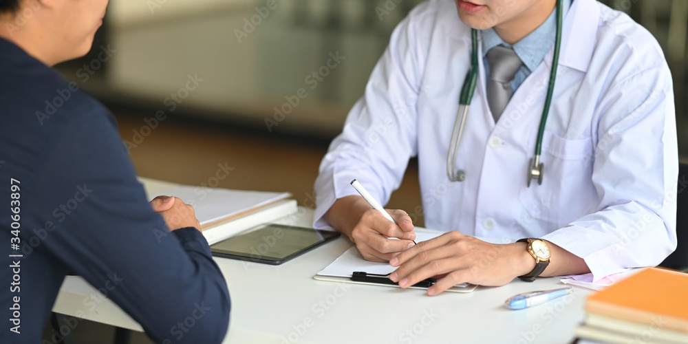 Fototapeta premium Cropped image of Young smart doctor diagnosing and giving advice to his patient while sitting together at the doctor working desk over comfortable clinic as background.