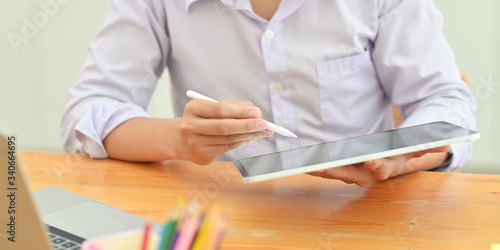 Cropped image of smart man in white shirt holding a computer tablet and stylus pen in his hands while sitting in front a computer laptop at the wooden working desk over living room as background.