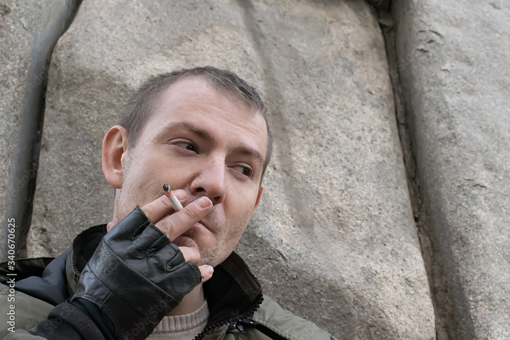 an adult guy, a man, with a cigarette and leather gloves is standing, hiding behind a stone obelisk and slyly looking, waiting for someone