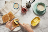 Female hands spreading butter on a toast. There are plate, coffee mocha, butter, marmalade, knife, cup with milk and napkin. From above.