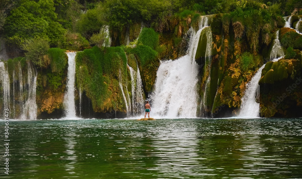 Fototapeta premium Waterfall in the nature with a young man flexing muscles. Green forest with a river. Near Plivice lakes in croatia