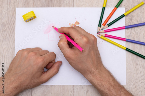 a man's hands draw with a pink pencil on a white sheet next to which are a yellow sharpener, shavings from a pencil and colored pencils scattered
