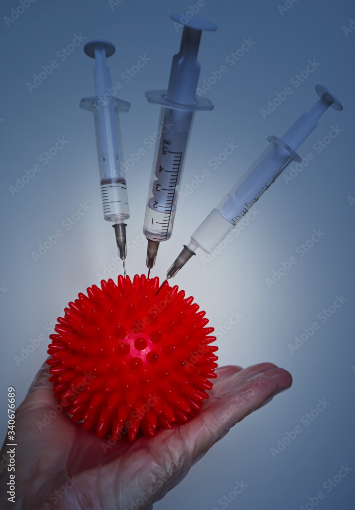 A hand in a white glove holds a Coronavirus and a syringes. Red symbol of virus COVID-19. Medical concept of vaccine, vaccination and fighting the epidemic and pandemic. A hand in a white glove holds a Coronavirus and a syringes. Red symbol of virus COVID-19. Medical concept of vaccine, vaccination and fighting the epidemic and pandemic.