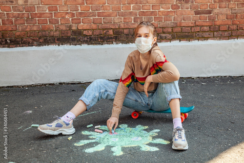 A teenage girl in a medical mask sits on a skateboard and draws a picture of the coronavirus with chalk on the pavement