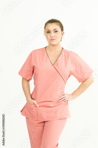 Attractive young Caucasian female healthcare worker poses in pink scrubs against plain background - serious with hands in pockets. photo