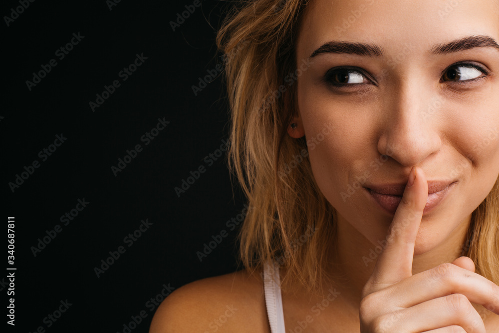 Young cheerful playful blonde woman posing on camera. Close up and cut view of girl holding finger on lips and look left. Hush symbol. Isolated over black background. Young cheerful playful blonde woman posing on camera. Close up and cut view of girl holding finger on lips and look left. Hush symbol. Isolated over black background.