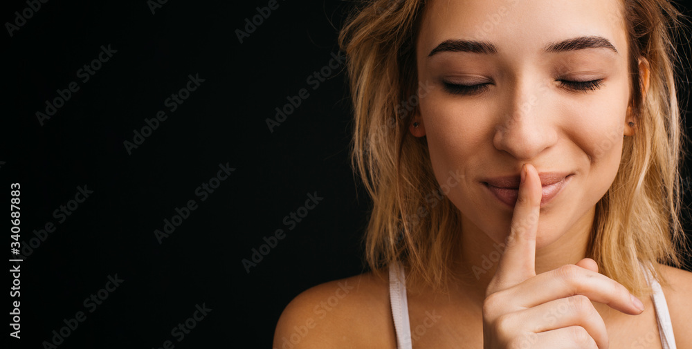 Naklejka premium Cheerful playful young woman posing on camera. Hold finger on lips and keep eyes closed. Hush symbol and quitet moment. Isolated over black background.