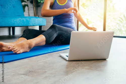20s young Asian woman in sportswear doing stretching exercises while watching yoga training class on computer laptop online. Healthy girl exercising in living room with sofa couch in the background. © twinsterphoto