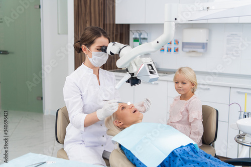 A blond woman having her teeth checked at the dentist s office  with her daughter waiting for her