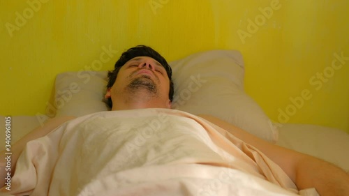 Portrait of sleeping caucasian bearded man at home in poor flat with torn off wallpaper , bottom view. He is breathing deep. He is lying on his back on pillow and covering blanket. Yellow background.