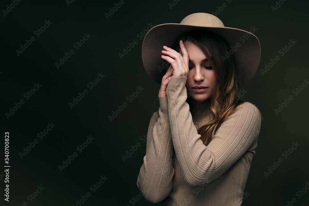 A young attractive blonde woman in a beige sweater, fedora hat and brown leather trousers poses on a dark green olive background. Studio portrait of a beautiful girl. Copy space. A young attractive blonde woman in a beige sweater, fedora hat and brown leather trousers poses on a dark green olive background. Studio portrait of a beautiful girl. Copy space.
