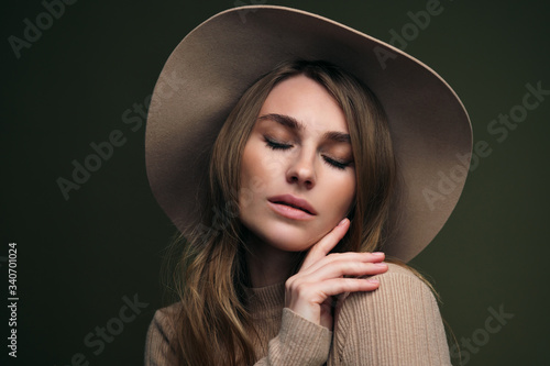 A young attractive blonde woman in a beige sweater, fedora hat and brown leather trousers poses on a dark green olive background. Studio portrait of a beautiful girl.