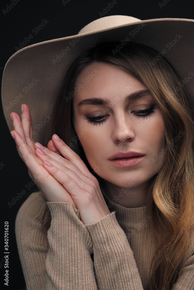 A young sensual sexy woman with a beautiful face and clear natural skin poses in the studio against a black background. Beige slinky sweater and hat with fields. A young sensual sexy woman with a beautiful face and clear natural skin poses in the studio against a black background. Beige slinky sweater and hat with fields.