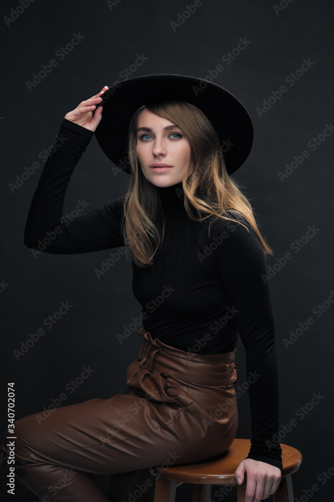 Portrait of young beautiful blond woman with light makeup and blue eyes touching her face. Dark background. Black tight sweater and hat with fields. Portrait of young beautiful blond woman with light makeup and blue eyes touching her face. Dark background. Black tight sweater and hat with fields.