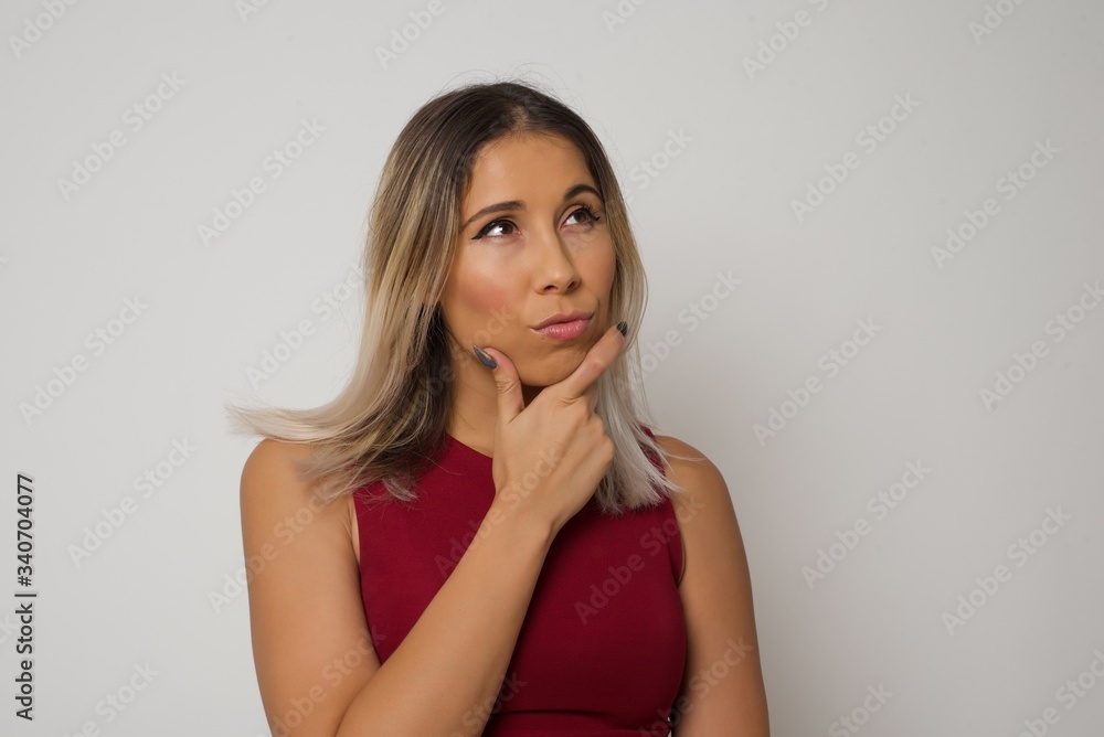 Isolated portrait of stylish young European woman with hand under chin and looking sideways with doubtful and skeptical expression, worry and doubt. Standing indoors over gray background. Isolated portrait of stylish young European woman with hand under chin and looking sideways with doubtful and skeptical expression, worry and doubt. Standing indoors over gray background.