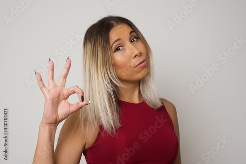 Glad attractive woman shows ok sign with hand as expresses approval, has cheerful expression, being optimistic. Standing against white wall.