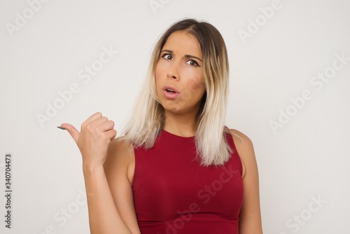 Horizontal shot of pretty young joyful shocked female points with thumb away, indicates something on blank wall isolated over white background. Check this out. Ethnicity concept.