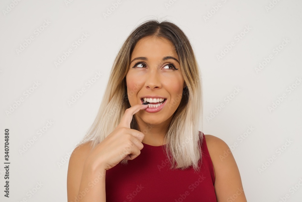 Dreamy female with thoughtful expression, looks to the camera, keeps hand near face, bitting a finger thinks about something pleasant, poses against gray wall. Dreamy female with thoughtful expression, looks to the camera, keeps hand near face, bitting a finger thinks about something pleasant, poses against gray wall.