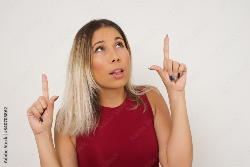 Horizontal shot of successful friendly looking young dark haired woman with bright make up exclaiming excitedly, pointing both index fingers up, indicating something shocking on blank copy space wall. Horizontal shot of successful friendly looking young dark haired woman with bright make up exclaiming excitedly, pointing both index fingers up, indicating something shocking on blank copy space wall.
