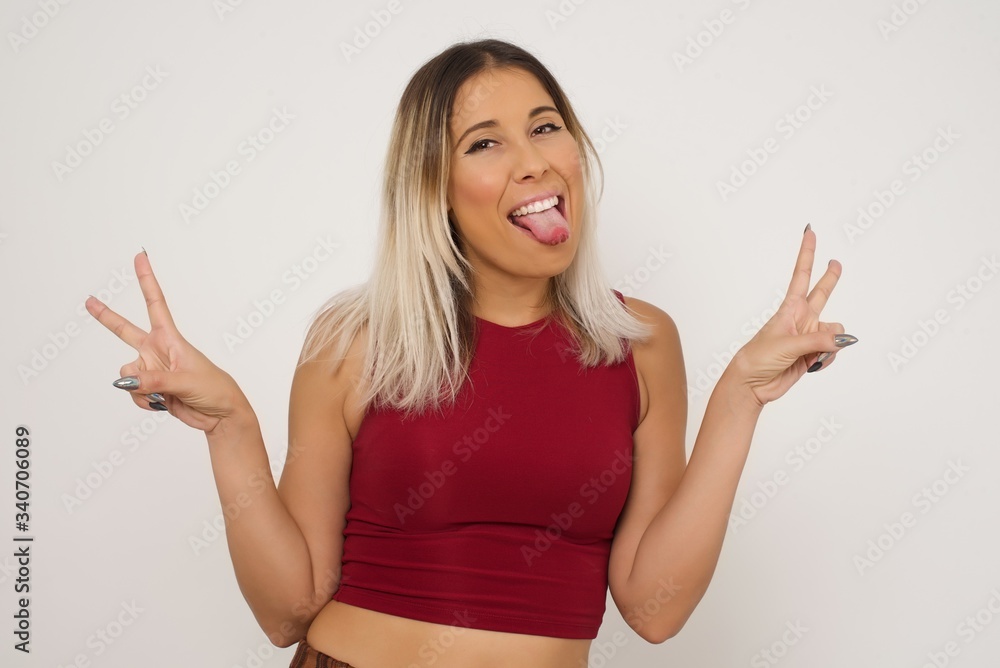 Fototapeta premium Indoor portrait of young caucasian female isolated on gray background with optimistic smile, showing peace or victory gesture with both hands, looking friendly. V sign.