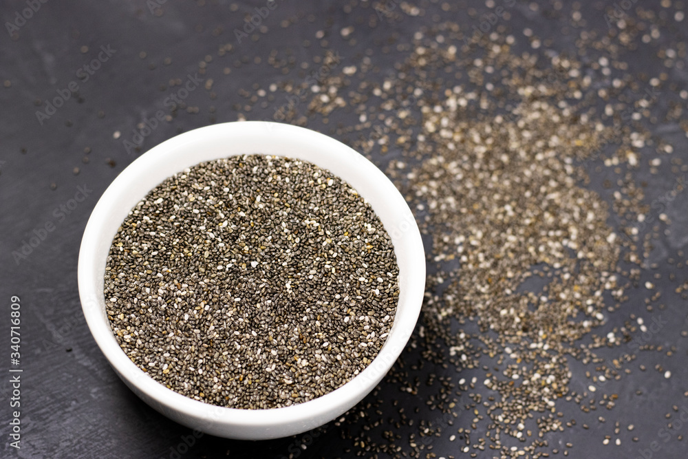 dark Chia seeds in a white bowl on a black stone background. small grains of Spanish sage, similar to beans, taste like a nut, gray-white-black color with a relief pattern. health product dark Chia seeds in a white bowl on a black stone background. small grains of Spanish sage, similar to beans, taste like a nut, gray-white-black color with a relief pattern. health product