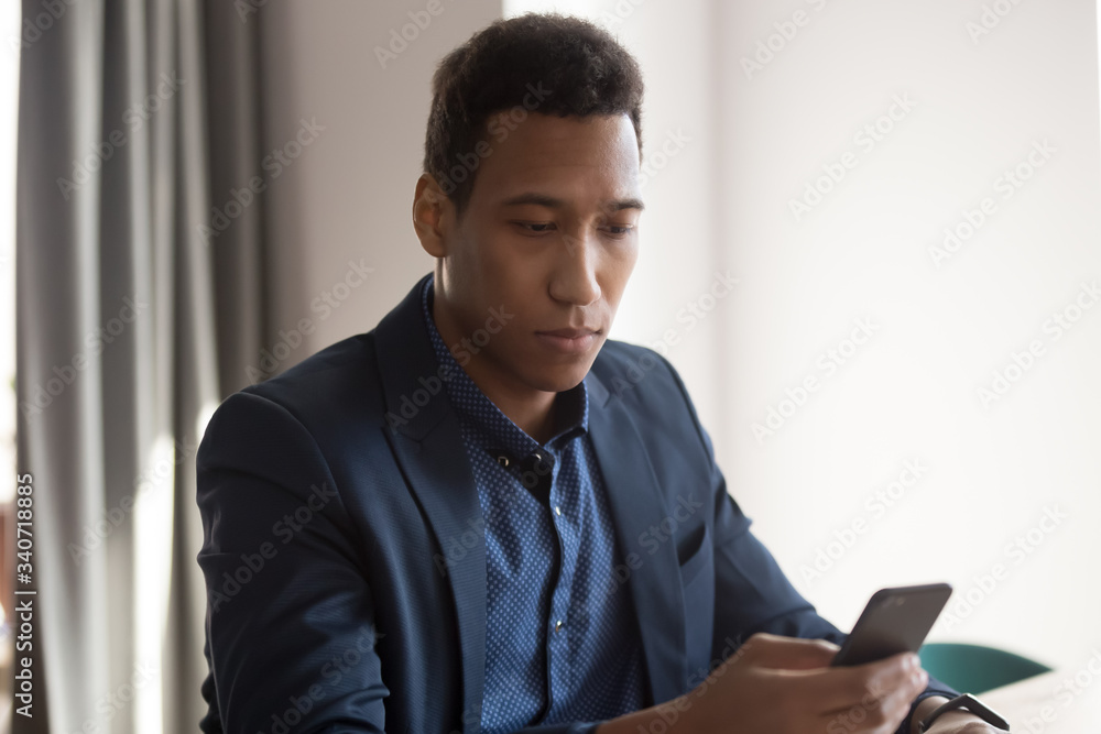 Serious mixed race businessman in suit holding smart phone looking at device screen read received message feels concerned sitting alone at workplace, employee browse internet, use business application Serious mixed race businessman in suit holding smart phone looking at device screen read received message feels concerned sitting alone at workplace, employee browse internet, use business application