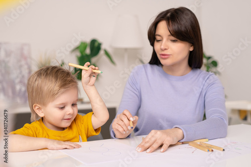 Cute little boy with crayon keeping hand raised over desk while drawing with mom