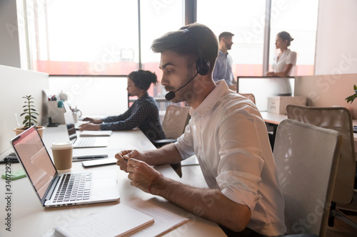 In shared room working employees, focus on call center agent wearing headset talking using computer consulting client online, operator telemarketer helping to customer via internet in office concept photo