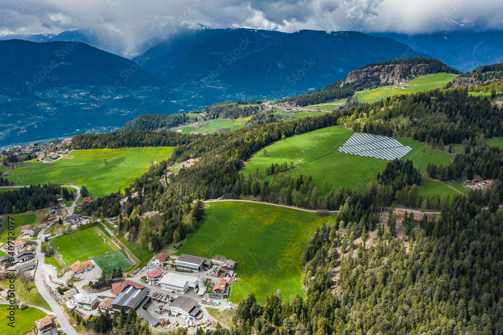Fototapeta premium Aerial view of farm of solar batteries on green slopes of the mountains of Italy, Trentino, huge clouds over a valley, roofs of houses of settlements, green meadows, a clear energy, energy of the sun