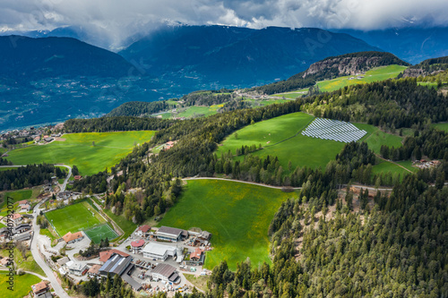 Aerial view of farm of solar batteries on green slopes of the mountains of Italy, Trentino, huge clouds over a valley, roofs of houses of settlements, green meadows, a clear energy, energy of the sun