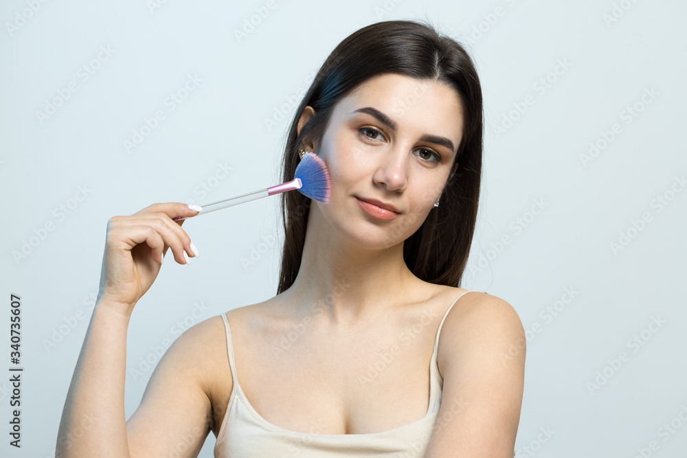 Close-up of a young girl in a light top on a white background making a facial make-up. A pretty woman holds a cosmetic brush near her face and smiles. Close-up of a young girl in a light top on a white background making a facial make-up. A pretty woman holds a cosmetic brush near her face and smiles.