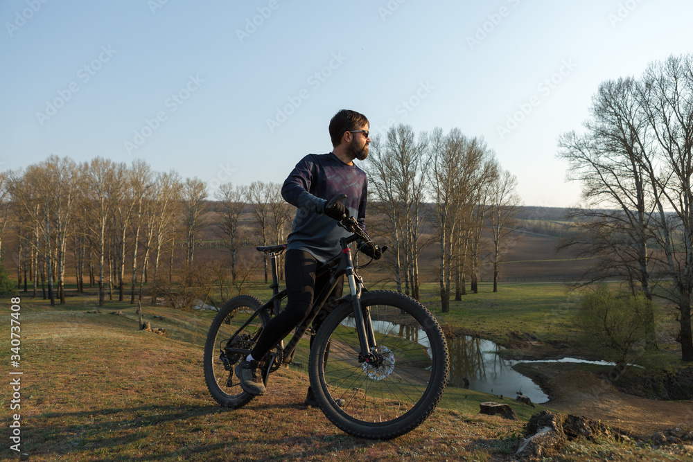 Cyclist in shorts and jersey on a modern carbon hardtail bike with an air suspension fork standing on a cliff against the background of fresh green spring forest