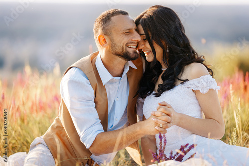 Happy beautiful couple in love in a flowering field on a hill laugh, smile, have fun and joke. Stylish wedding photo session in rustic style. Romantic portrait of the bride and groom on their wedding
