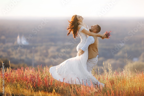 A loving groom circles the bride in his arms on the field in nature. Romantic portrait of the bride and groom on a wedding day in a rustic style.