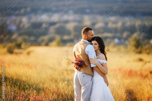 Young beautiful couple in a flowering field on a hill hug each other at sunset. Rustic wedding photoshooting. Romantic sensual portrait of the bride and groom on their wedding day