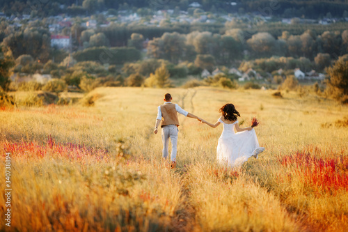 Young beautiful couple in love in a flowering field on a hill run along the road at sunset. Stylish wedding photo session in rustic style. Romantic portrait of the bride and groom on their wedding day © Саша Тихова