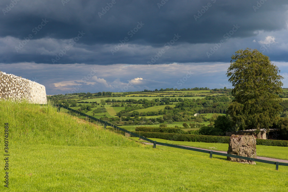The dazzling white quartz stone façade of Newgrange megalithic passage tomb contrasts with the lush green rolling hills of the Irish countryside and dark storm clouds on a summers day.