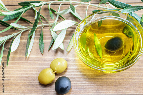 Olive oil in glass transparent bowl, black and green olives and olive tree branches in sunlight on the wooden background.