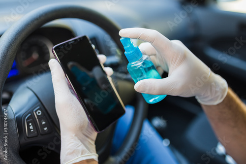 Driver in gloves using disinfectant spray bottle to clean mobile phone screen. Car interior.