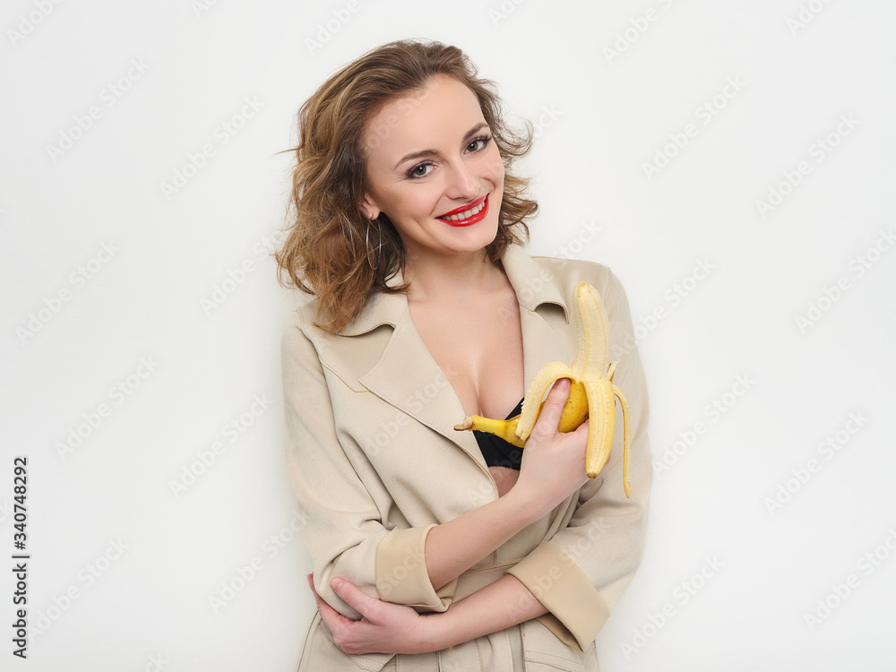 Sexy woman with red lips and curly hairdo in short coat and black bra playfully posing eating banana on white background Sexy woman with red lips and curly hairdo in short coat and black bra playfully posing eating banana on white background