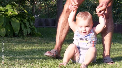 Grandfather teaches the toddler boy to walk. Baby boy making his first steps. The kid tries to walk barefoot on the grass in the summer in the back yard and holds dad's hands. Front view.