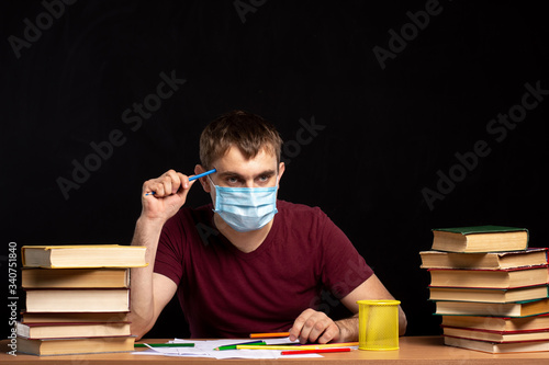 young man in a medical mask sits at a table with books on a background of a black wall. holding a pencil in his hand near his face. quarantine work. pensive look.