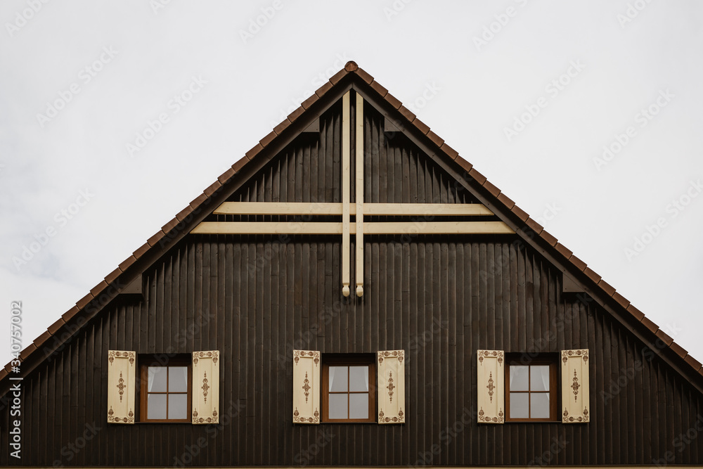 Gable roof of cottage with cloudy sky in background.There are 3 glass windows with wooden decorated shutters. Gable is made of wooden planks with dominant wooden cross at its top.