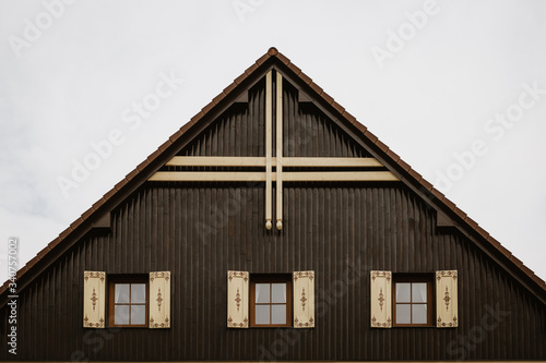 Gable roof of cottage with cloudy sky in background.There are 3 glass windows with wooden decorated shutters. Gable is made of wooden planks with dominant wooden cross at its top. © Zdena Venclik