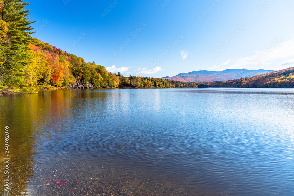 Majestic mountain lake surrounded by deciduous forest at the peack of fall foliage on a sunny autumn morning. Reflection in water.