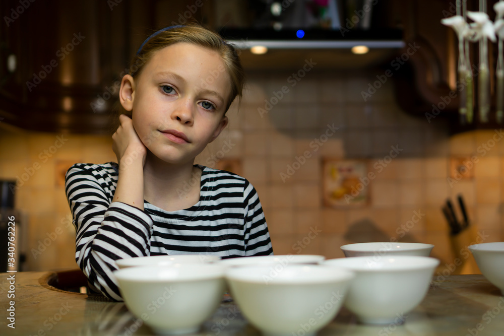 Cute caucasian blond child girl in a striped dress does not know what to cook sitting at cosy home kitchen table with many empty white plates bowls Cute caucasian blond child girl in a striped dress does not know what to cook sitting at cosy home kitchen table with many empty white plates bowls
