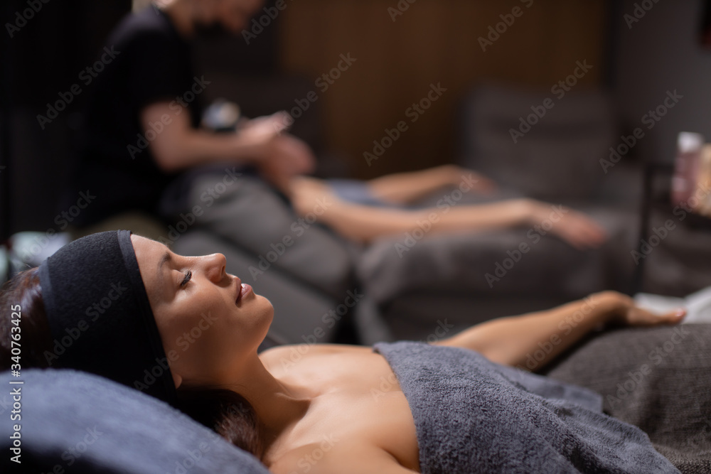 side view on calm relaxed caucasian woman lying in wellbeing salon before skin treatment, woman get face lift massage in the background side view on calm relaxed caucasian woman lying in wellbeing salon before skin treatment, woman get face lift massage in the background